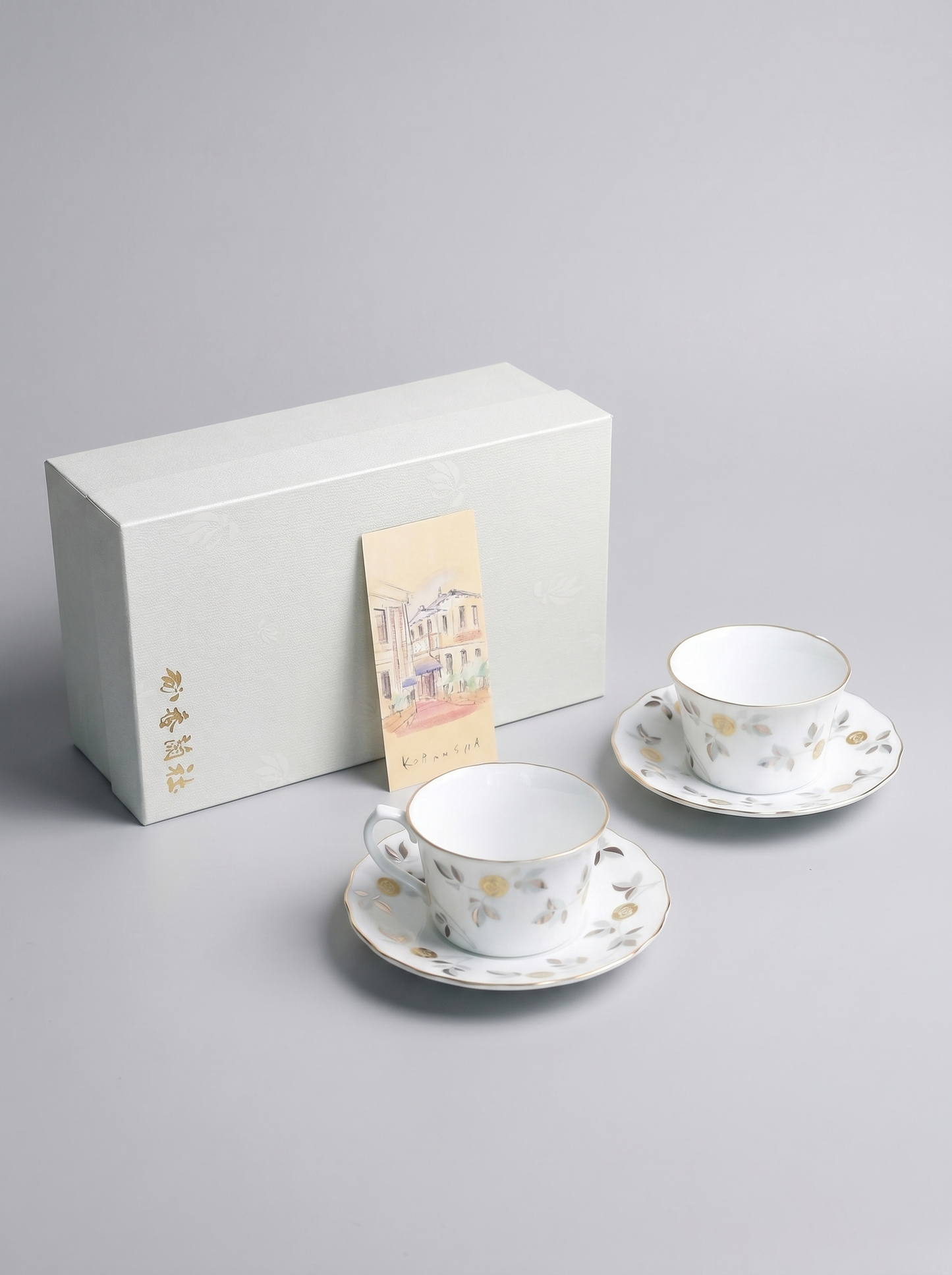 Two ceramic tea cups with saucers and a white box on a gray background