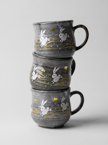 Stack of three gray mugs with rabbit illustrations on a light gray background