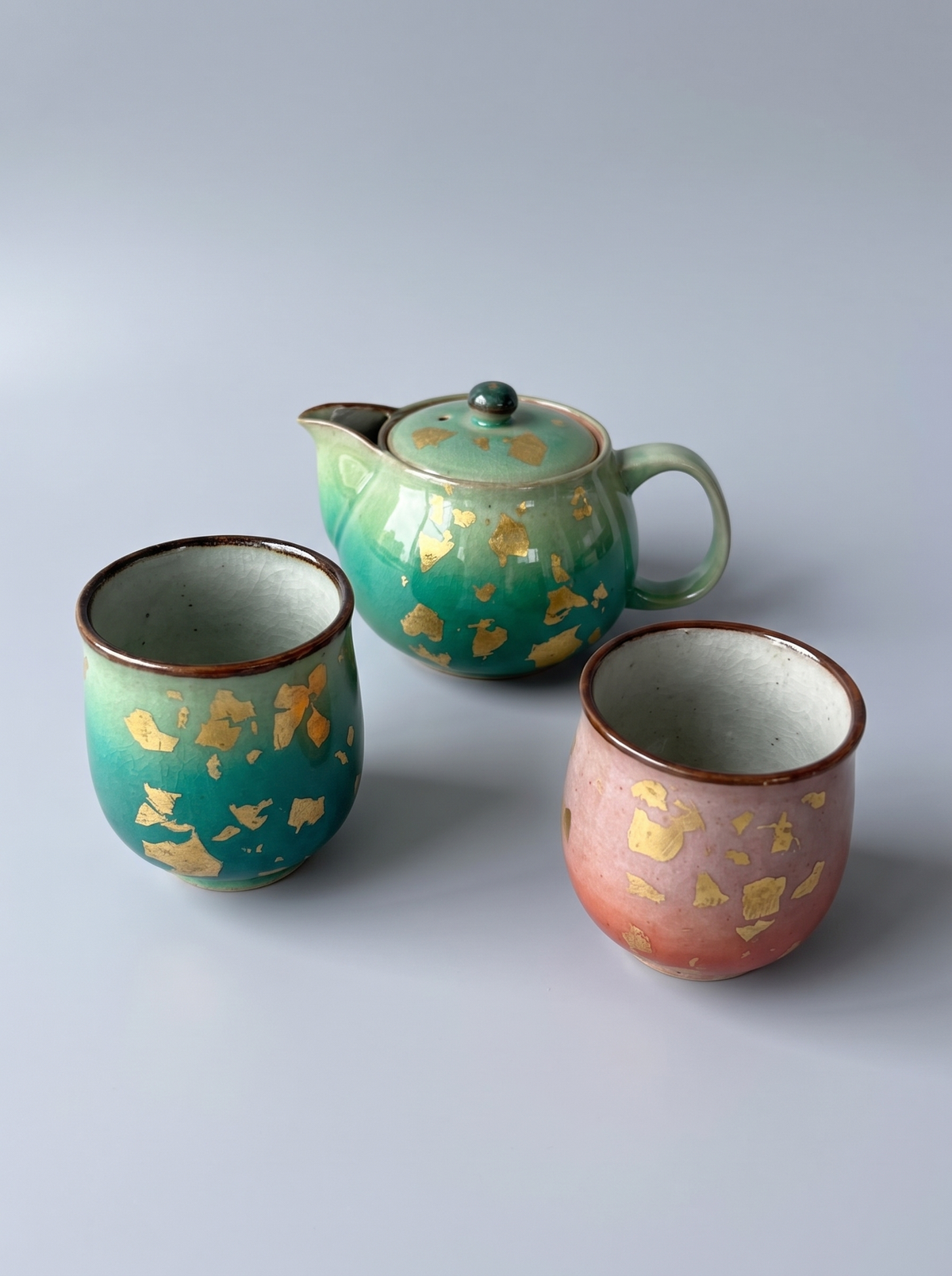 Three handmade ceramic teaware pieces on a light gray background: one lidded teapot and two cups. The teapot is teal-green with scattered gold leaf fragments, and the left cup is a similar teal-green with gold leaf fragments and a brown rim.