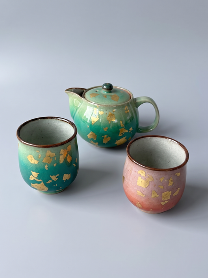 Three handmade ceramic teaware pieces on a light gray background: one lidded teapot and two cups. The teapot is teal-green with scattered gold leaf fragments, and the left cup is a similar teal-green with gold leaf fragments and a brown rim.