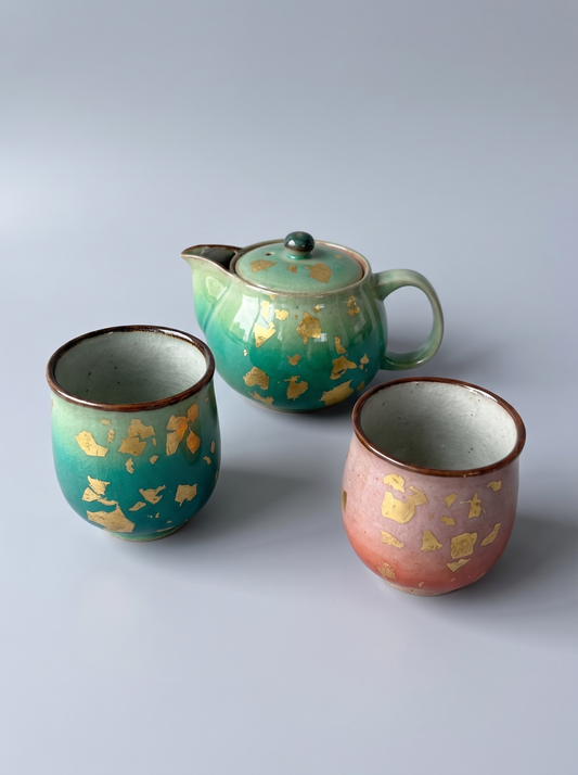 Three handmade ceramic teaware pieces on a light gray background: one lidded teapot and two cups. The teapot is teal-green with scattered gold leaf fragments, and the left cup is a similar teal-green with gold leaf fragments and a brown rim.