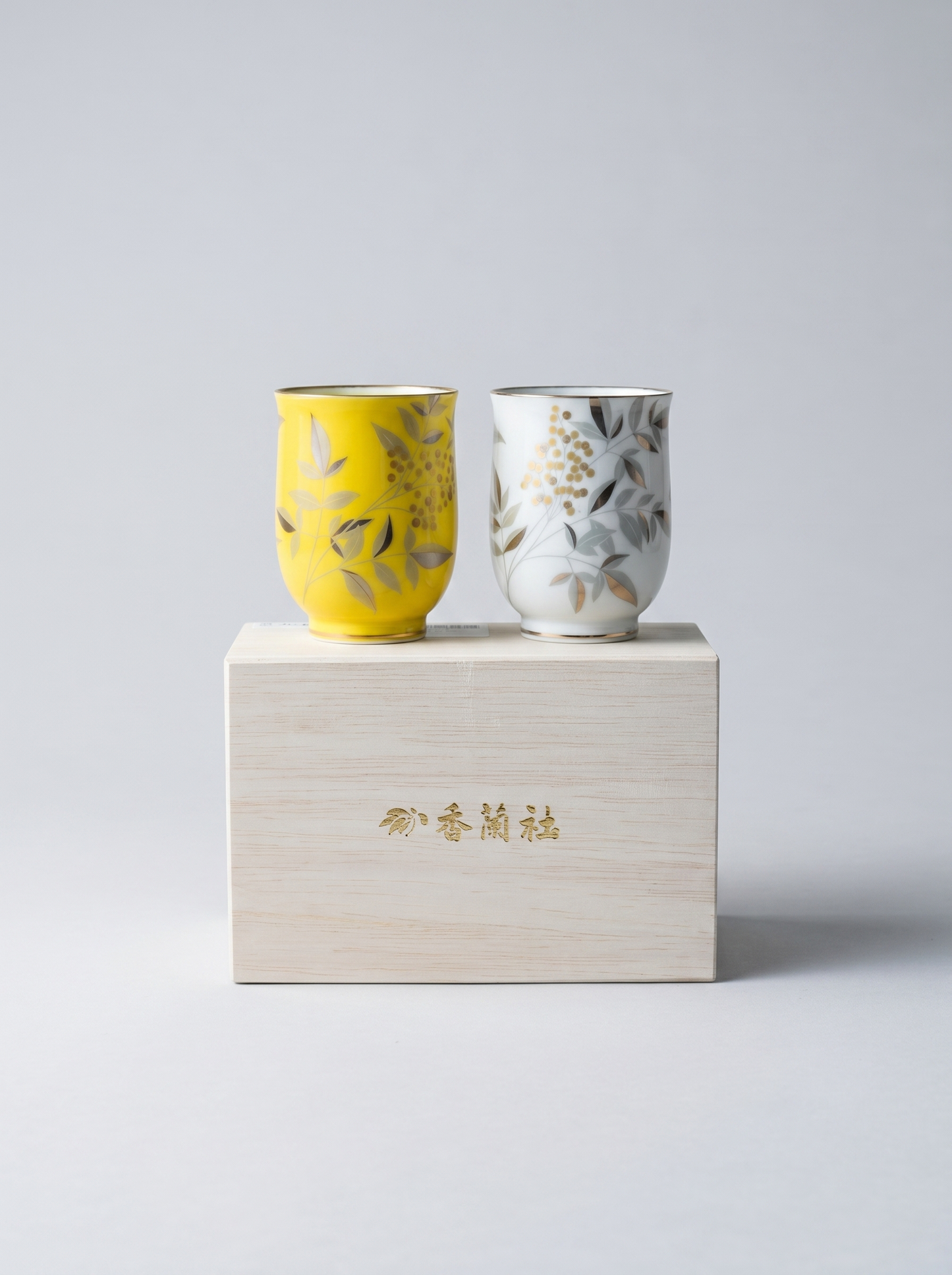 Two ceramic cups, one yellow and one white with floral patterns, on a gift box against a light gray background.