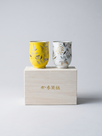 Two ceramic cups, one yellow and one white with floral patterns, on a gift box against a light gray background.
