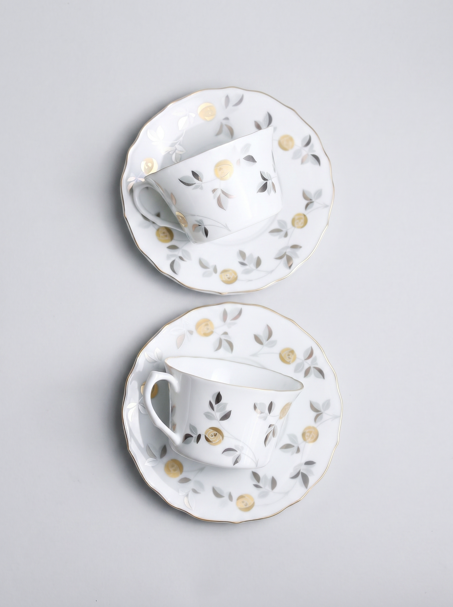 Two white ceramic tea cups with saucers featuring gold accents and floral patterns on a light gray background.