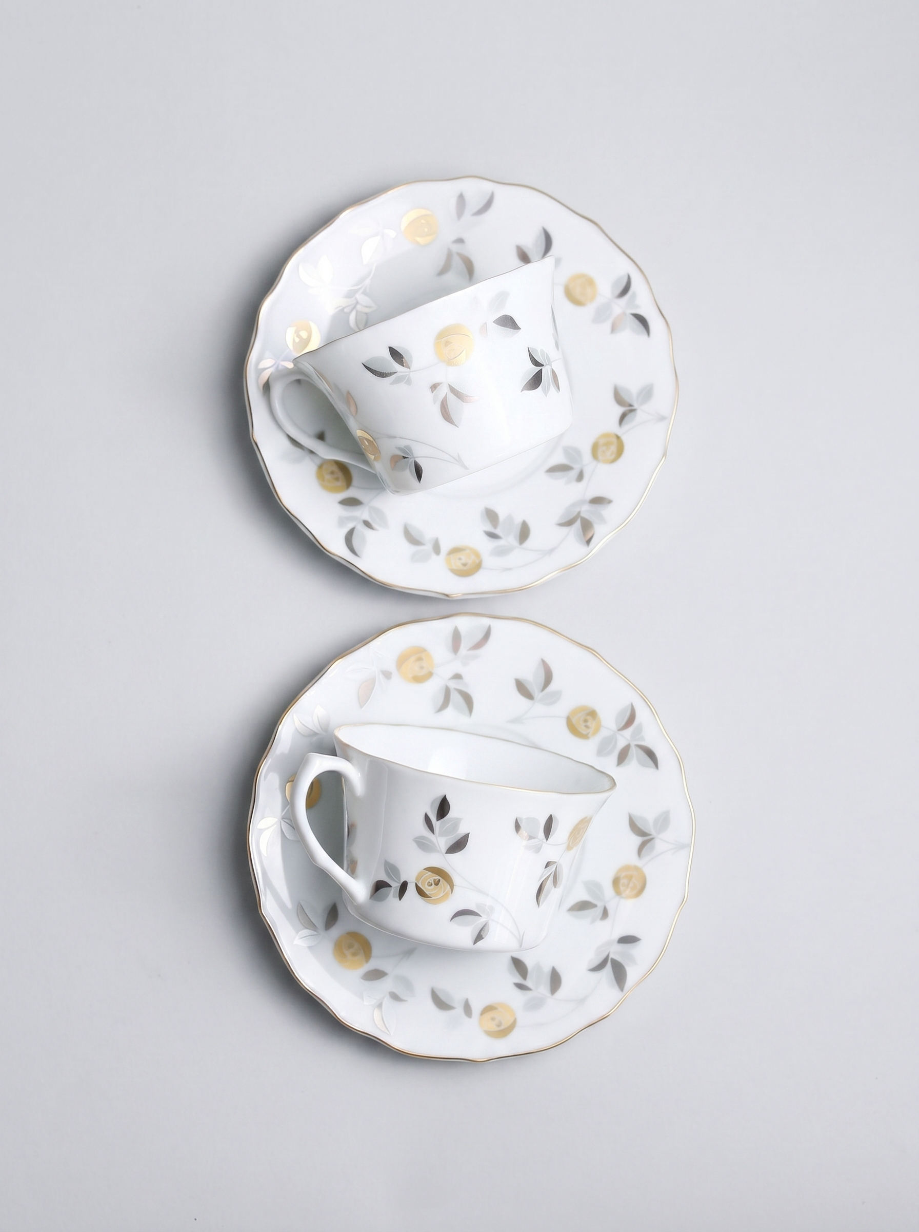 Two white ceramic tea cups with saucers featuring gold accents and floral patterns on a light gray background.