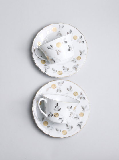 Two white ceramic tea cups with saucers featuring gold accents and floral patterns on a light gray background.