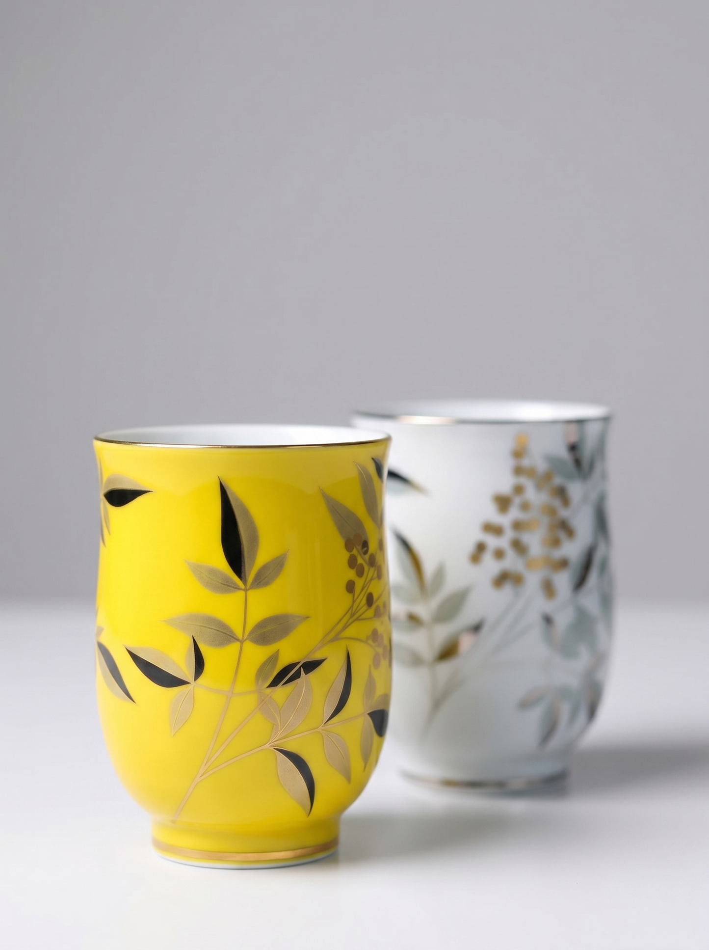 Two ceramic cups, one yellow with golden leaf design and the other white with gold leaf design, on a light gray background.