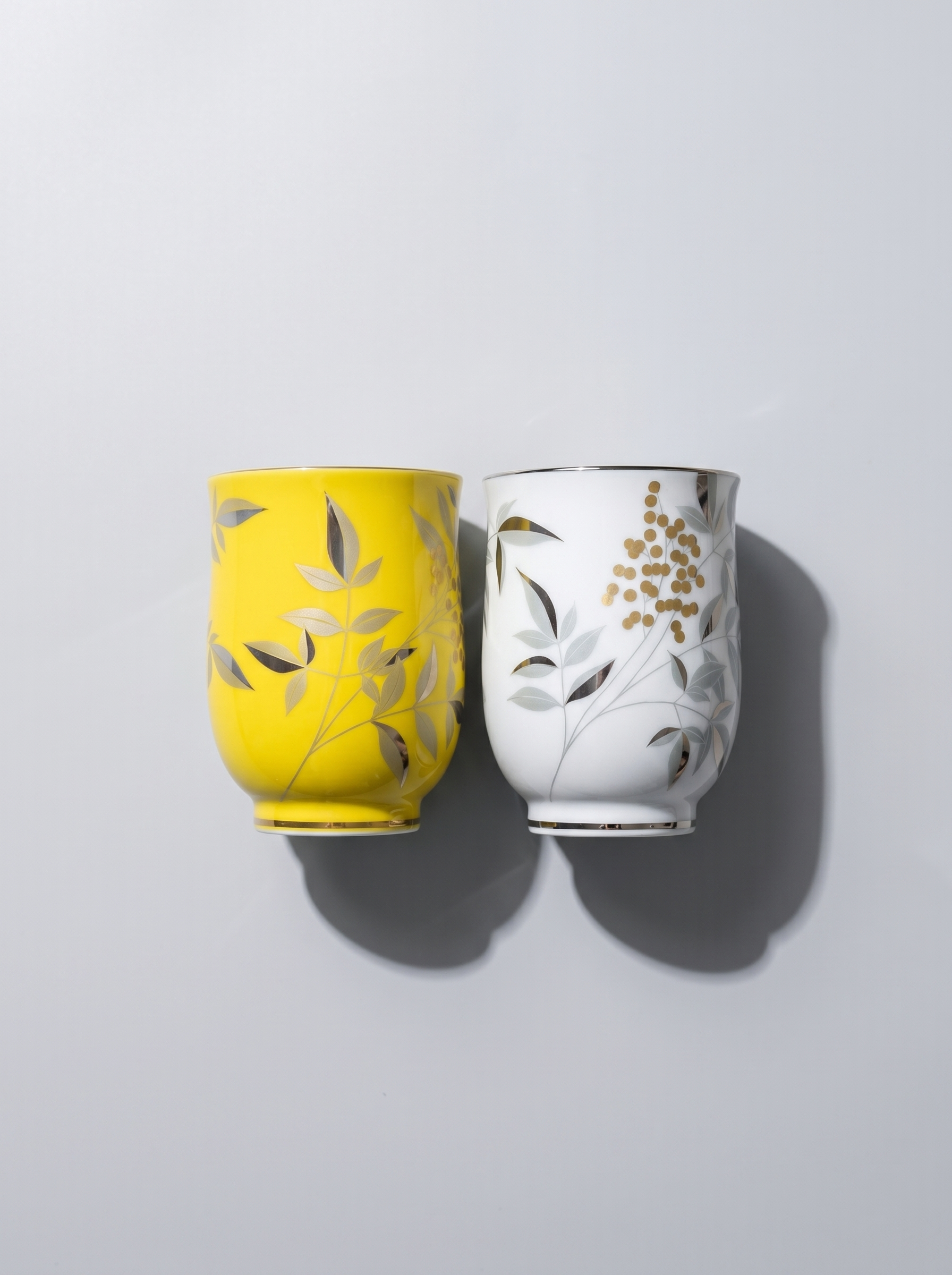 Two ceramic cups, one yellow and one white, with leaf patterns on a light gray background.