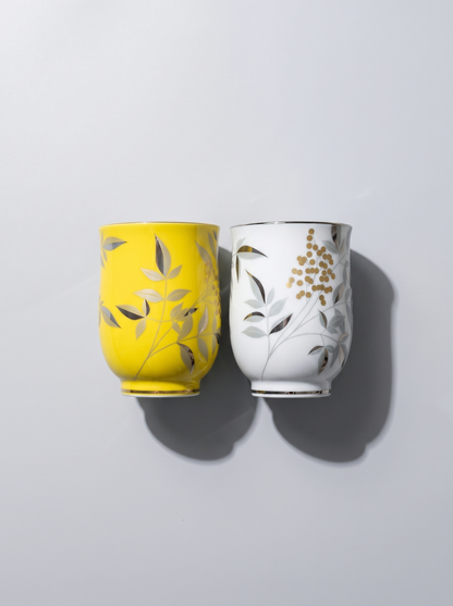 Two ceramic cups, one yellow and one white, with leaf patterns on a light gray background.