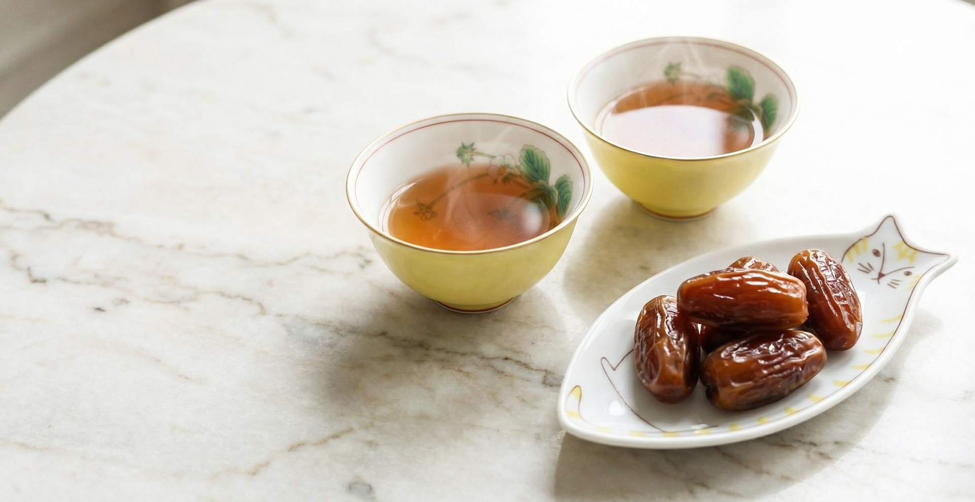 Two Japanese porcelain cups of tea and a cat-shaped sauce plate of dates on a marble surface