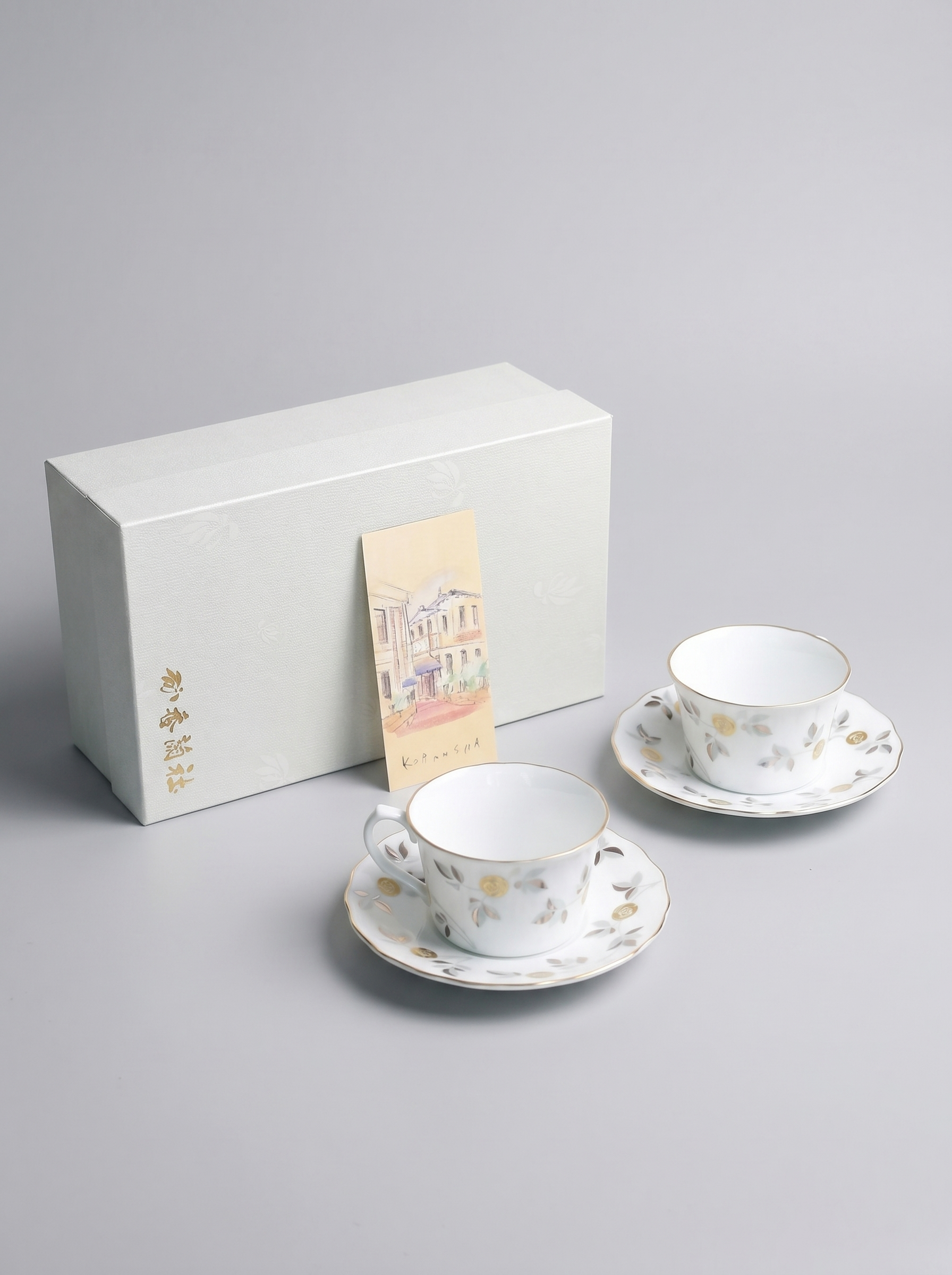 Two ceramic tea cups with saucers and a white box on a gray background