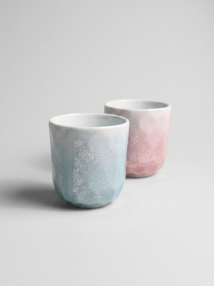 Two ceramic cups with ginsai finish, one light blue and one pink, on a gray background