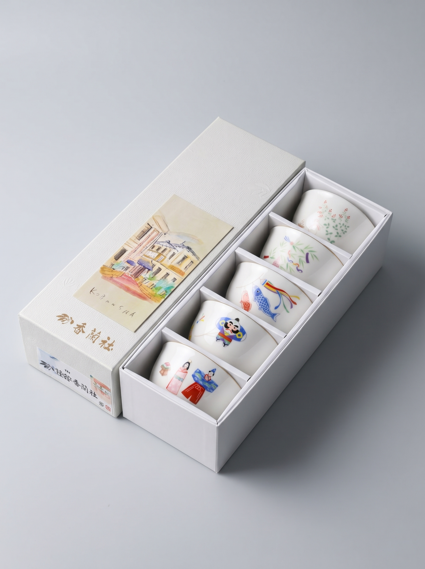 Set of five small porcelain cups with colorful designs in a white Japanese gift box on a light gray background