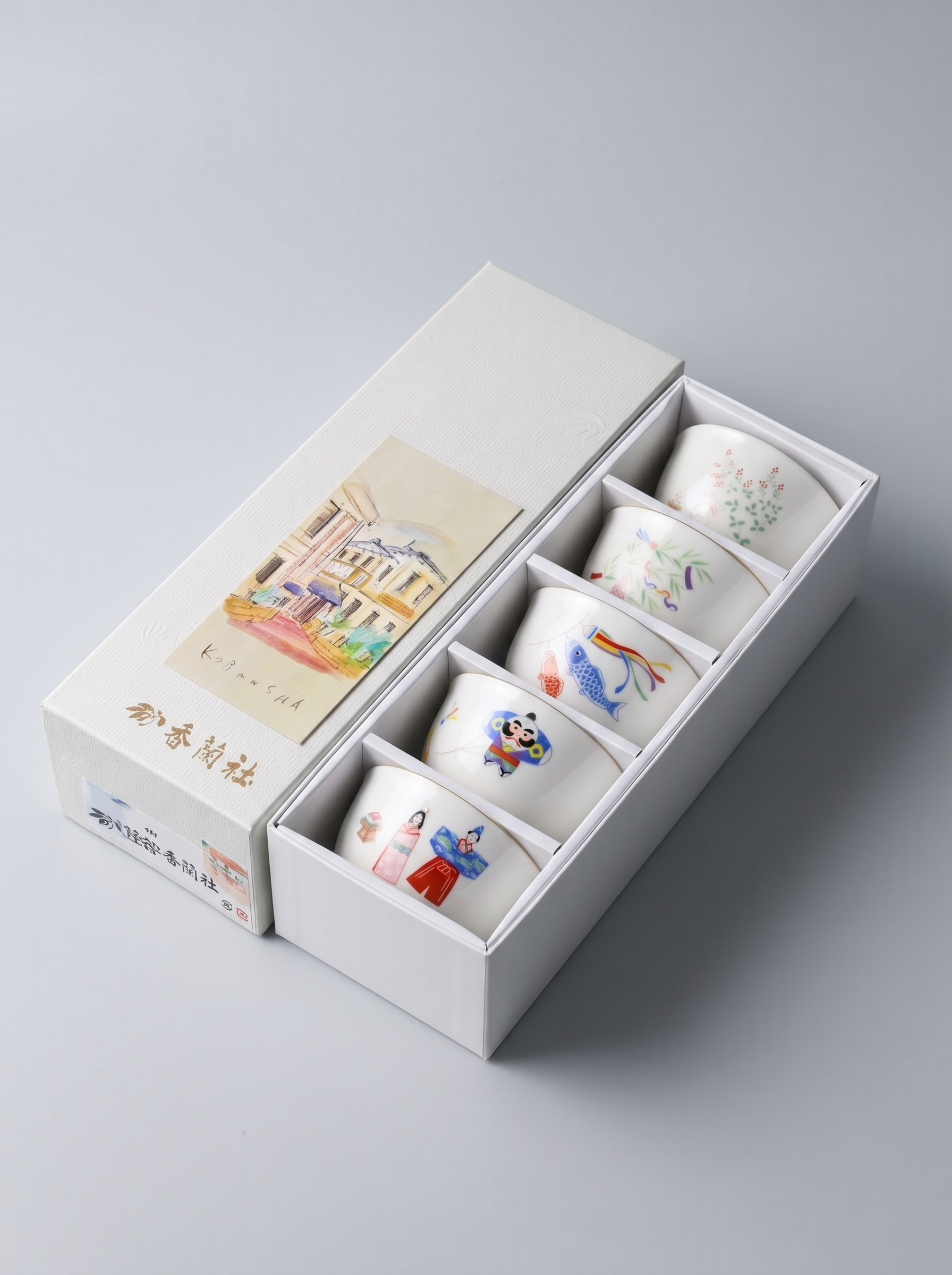 Set of five small porcelain cups with colorful designs in a white Japanese gift box on a light gray background
