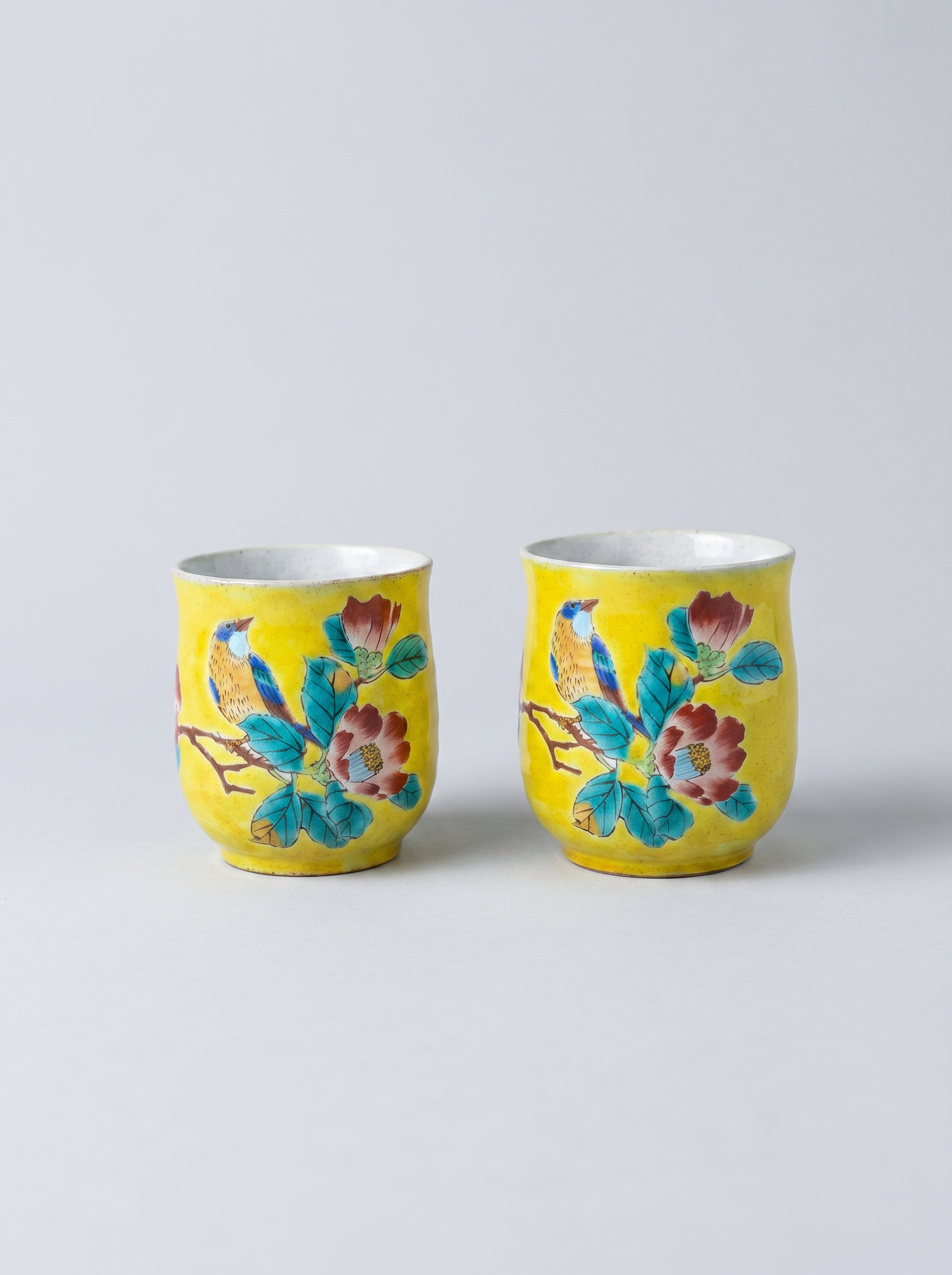 Two yellow Japanese ceramic cups with floral designs in yoshidaya style on a light gray background