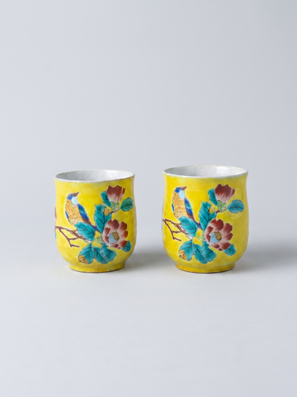 Two yellow Japanese ceramic cups with floral designs in yoshidaya style on a light gray background
