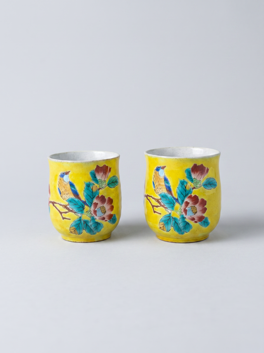 Two yellow Japanese ceramic cups with floral designs in yoshidaya style on a light gray background
