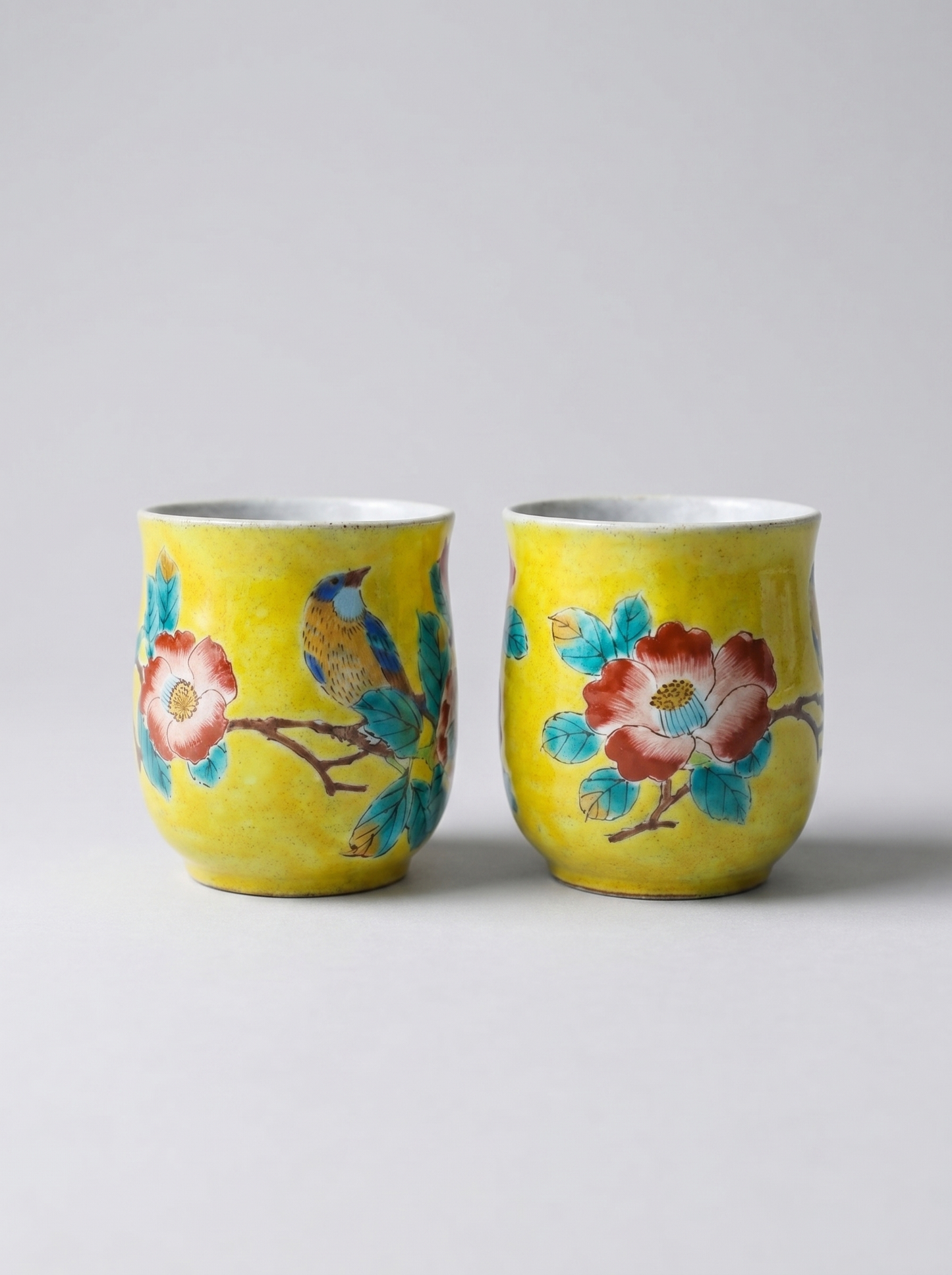 Two yellow ceramic cups with floral and bird designs on a light gray background