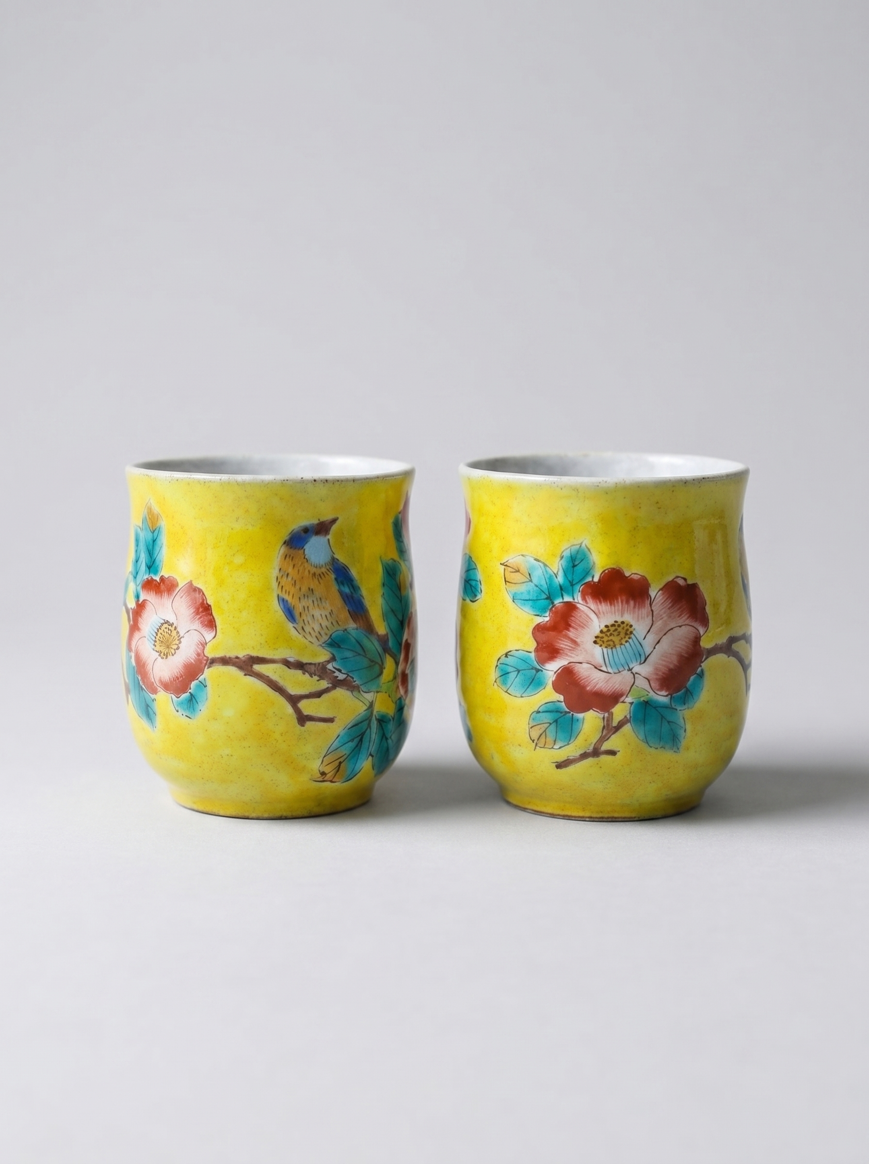 Two yellow ceramic cups with floral and bird designs on a light gray background