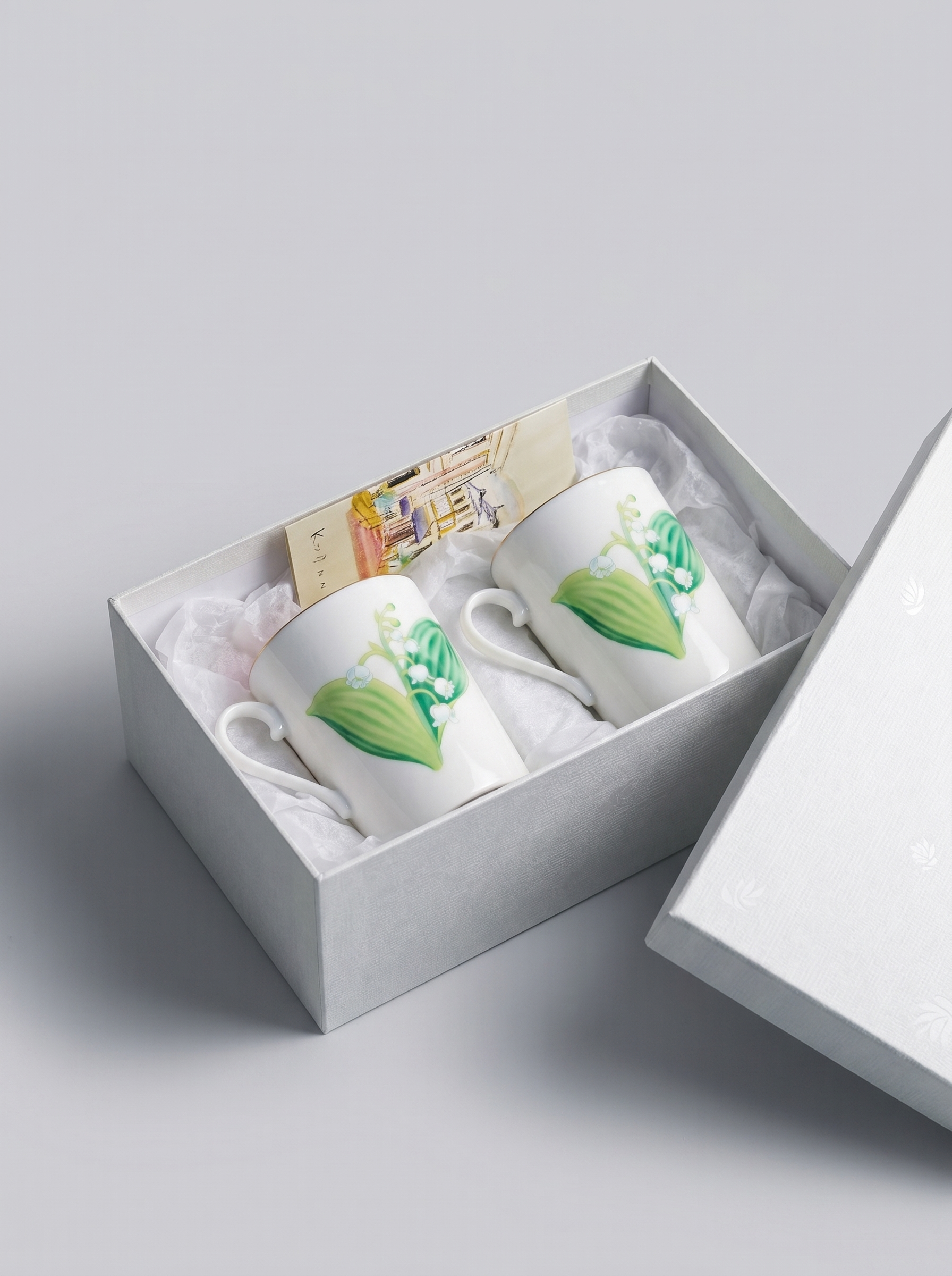 Two white porcelain teacups with lily of the valley designs in a gift box on a light gray background