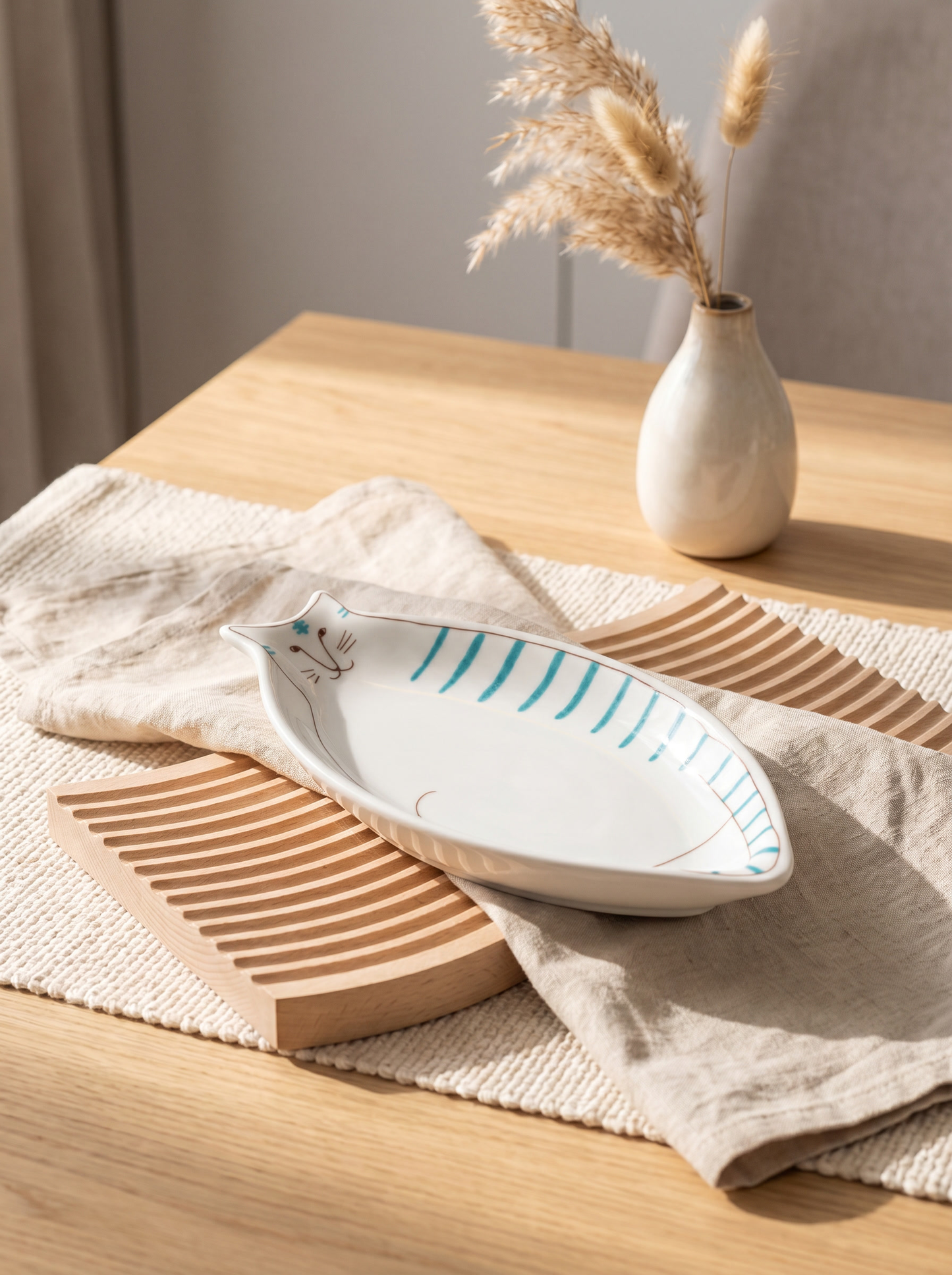 White ceramic cat-shaped dinner plate with blue stripes on a wooden table with a vase and dried plants.