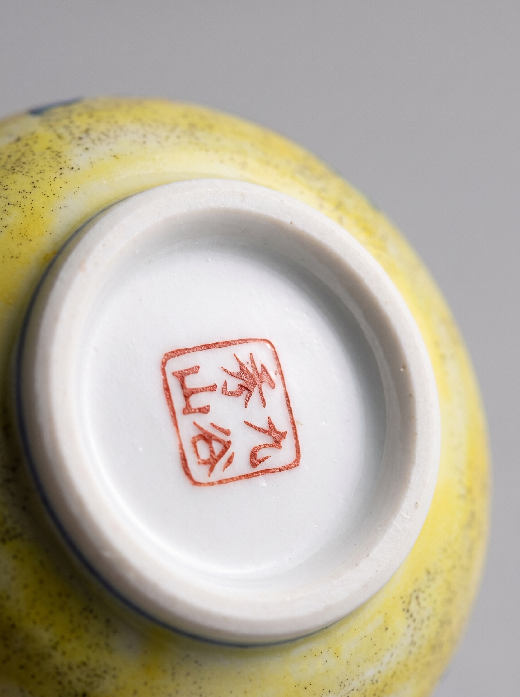 Close-up of a ceramic bowl with a yellow glaze and red seal mark on a gray background