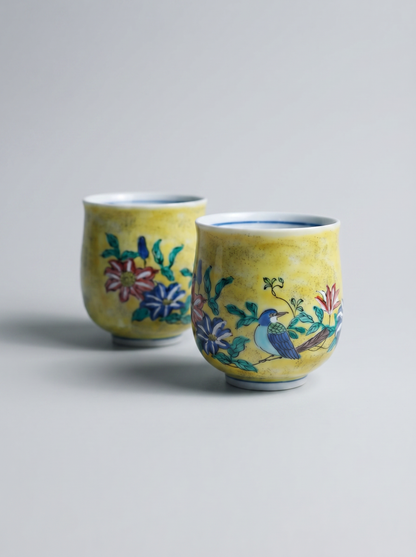 Two yellow ceramic cups with floral and bird designs on a gray background